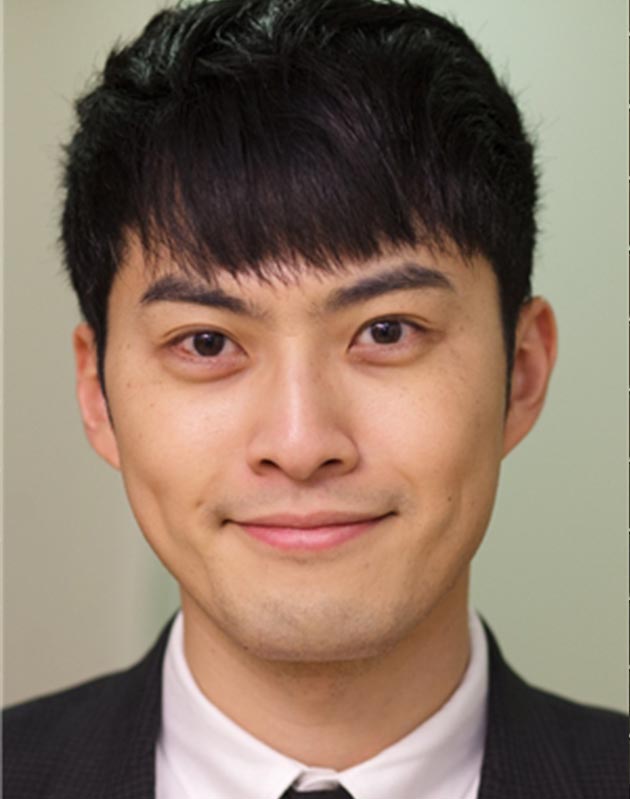 The image shows a man with short hair smiling at the camera while wearing a dark suit and tie.