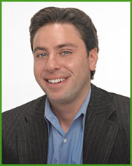 The image shows a man smiling at the camera with his head slightly tilted, wearing a dark suit with a blue shirt underneath, set against a plain background.