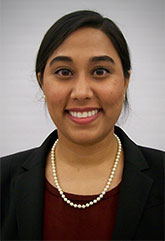 The image shows a smiling individual wearing a dark blazer over a lighter shirt, with a pearl necklace and a neutral expression.