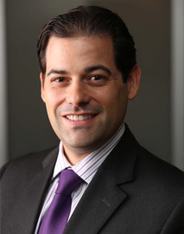 The image shows a man wearing a suit, smiling at the camera, with his head slightly tilted.