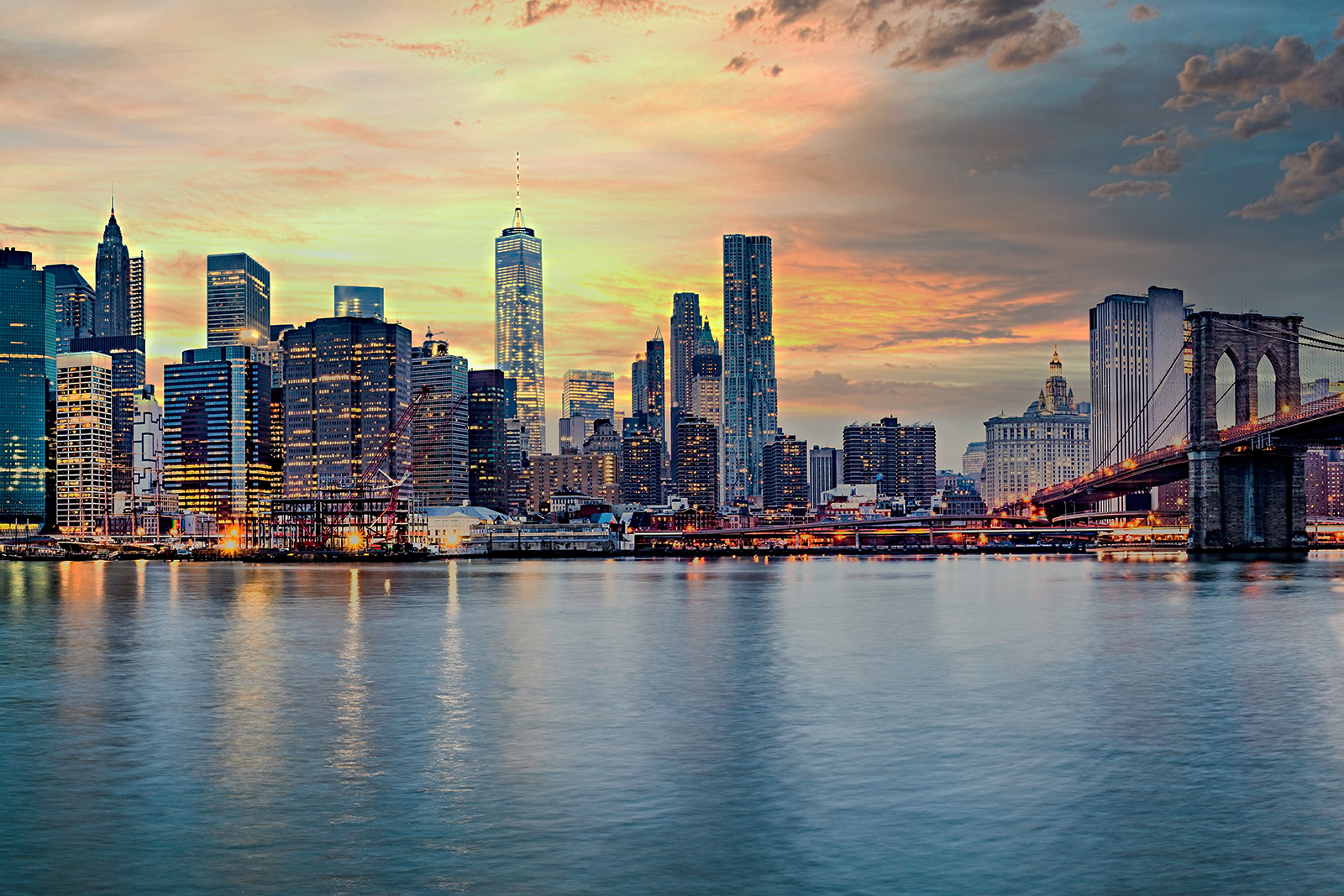 The image shows a city skyline with the Manhattan Bridge visible over a river at sunset, with the city s lights reflecting on the water surface.