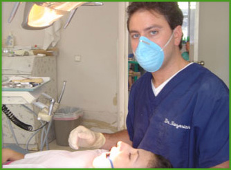 The image shows a man wearing medical scrubs and a surgical mask standing at a dental workstation with a patient lying on a dental chair, and he appears to be performing dental surgery or an examination.