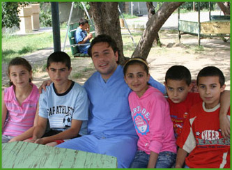 The image depicts a group of people posing together outdoors, with a man standing among them who appears to be in a medical uniform, smiling at the camera.