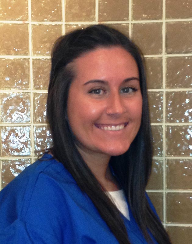 This is a photograph of a woman standing against a tiled wall. She has long dark hair, wears a blue uniform with a light-colored collar, and has a smile on her face. The woman appears to be in a professional setting, possibly within a healthcare facility given the attire and background.