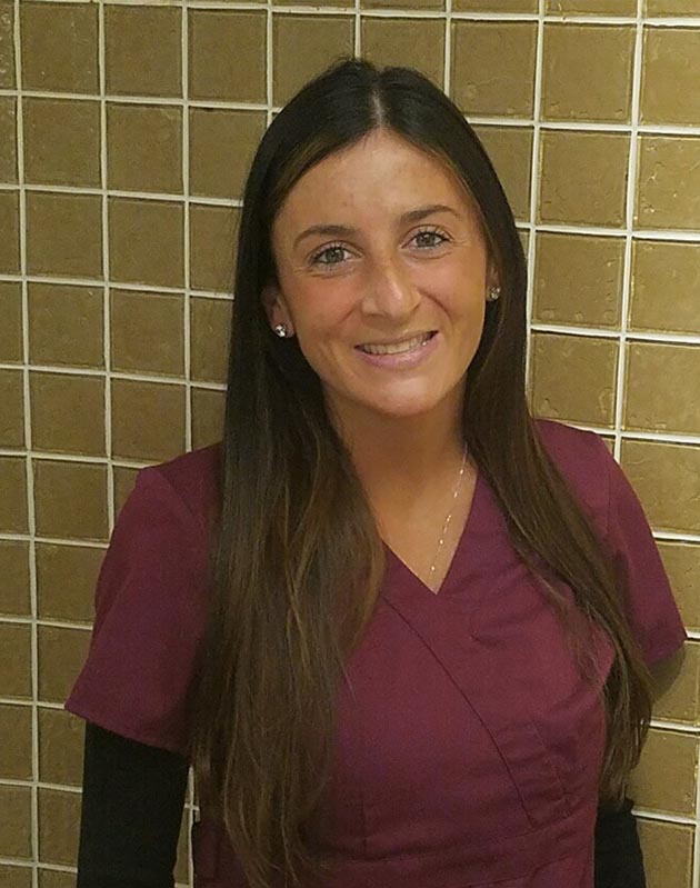 A woman wearing a maroon scrub top stands against a beige tiled wall.