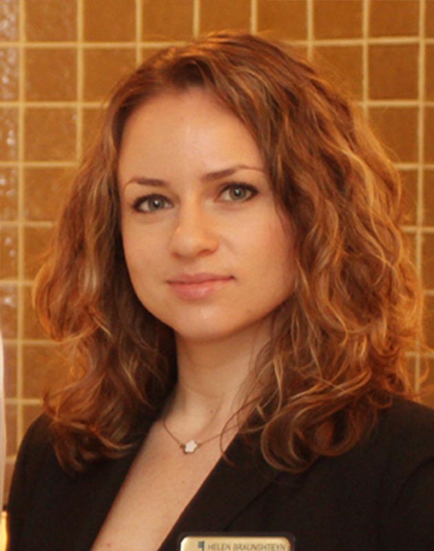 The image shows a woman with short hair, wearing a dark blazer, standing in front of a wall with tiles, posing for the photograph.