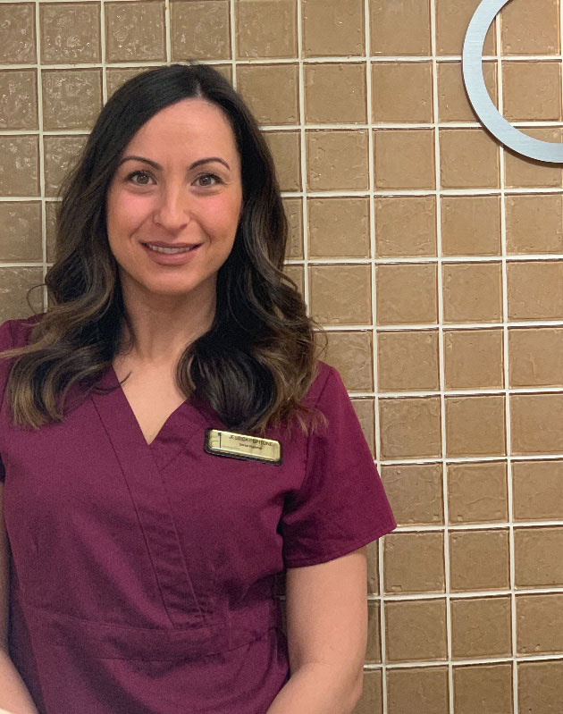 The image shows a woman standing against a wall with tiles, wearing a maroon scrub top and smiling at the camera.