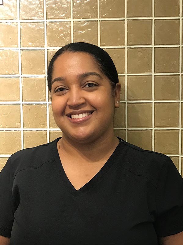 The image shows a woman wearing a black shirt with a white collar and a smile on her face, standing against a tiled wall.