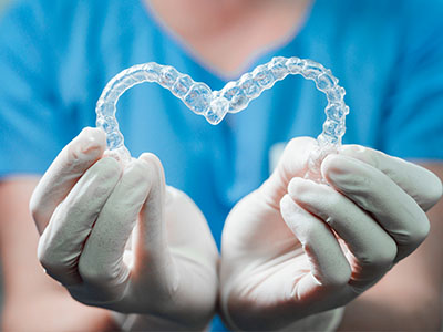 The image shows a pair of clear braces held by gloved hands against a backdrop featuring a blue surgical gown, suggesting a dental or orthodontic theme related to oral health care.
