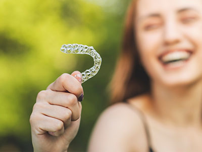 A smiling woman holds up a clear plastic dental retainer with her right hand.