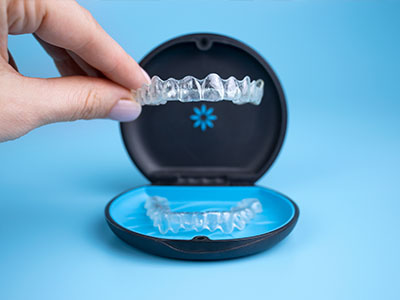 The image shows a hand holding an open case containing a set of clear dental aligners, with one aligner partially inserted into a mouth model for demonstration purposes.