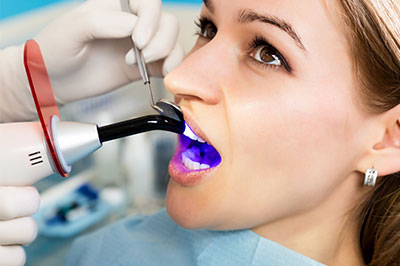 The image shows a woman seated in a dental chair with her mouth open, receiving dental care from a professional using a device to clean or examine her teeth.