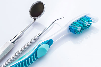 The image shows a collection of dental tools including a toothbrush with blue bristles, a pair of tweezers, and a dental mirror, all placed on a white surface against a light background.