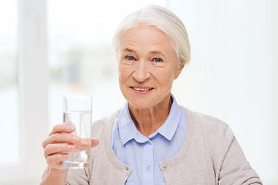 An elderly woman holding a glass of water, smiling slightly, against a blurred background.