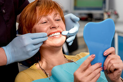 The image shows a woman sitting in a dental chair, smiling at the camera while holding a toothbrush-shaped object, with a dental professional seated behind her, performing a procedure on her teeth.