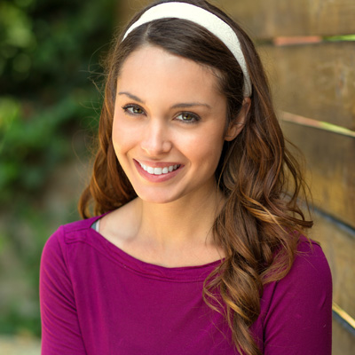 The image shows a smiling woman with long brown hair wearing a purple top and a headband, standing outdoors against a wooden fence.