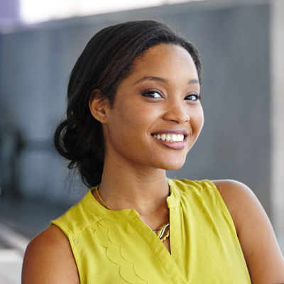 A woman with a warm smile, wearing a yellow blouse, stands confidently against a backdrop of a building wall.