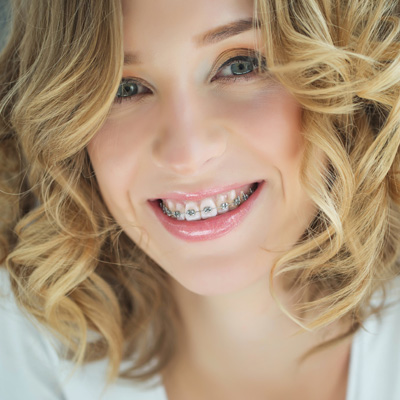 A smiling woman with braces, wearing a white top and curly hair, posing for a portrait with a soft focus background.