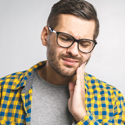 This is a photograph of a man with a beard wearing glasses and a plaid shirt, holding his hand to his mouth and looking downwards with an expression of concern or pain.