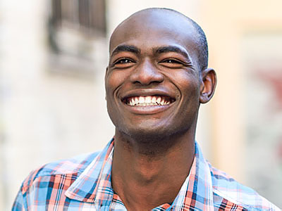 A smiling man with short hair, wearing a plaid shirt, stands against a wall with a window.